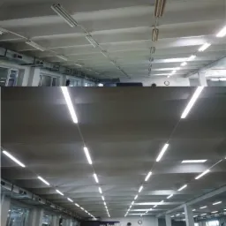 Rzędy lamp fluorescencyjnych zamontowane na suficie hali produkcyjnej, widoczne przewody zasilające, zmodernizowane oświetlenie w obiekcie przemysłowym.