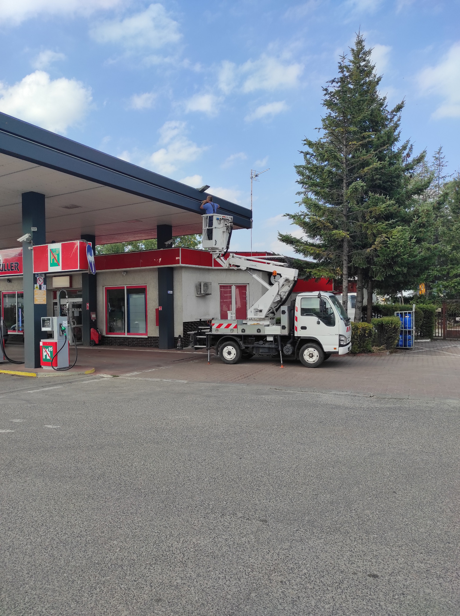 Pracownik na podnośniku koszowym przy stacji benzynowej, prawdopodobnie wykonuje prace elektryczne związane z oświetleniem zewnętrznym. Widoczny budynek stacji, dystrybutory paliwa i wysoka sosna...