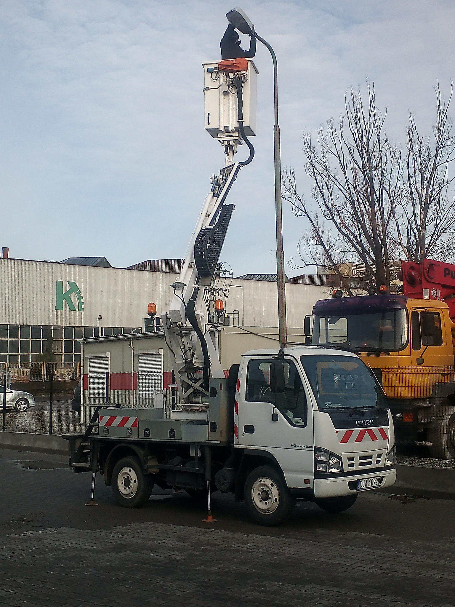 Pracownik w podnośniku koszowym, naprawiający latarnię uliczną na tle budynku z logo i zaparkowanych ciężarówek.