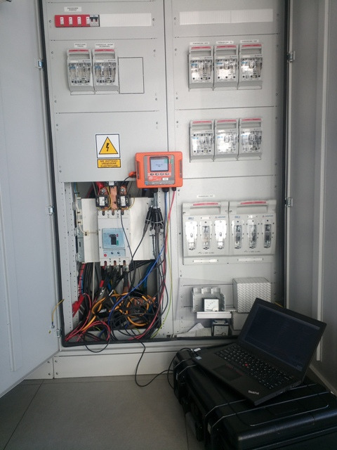 Otwarta szafa elektryczna z widocznymi bezpiecznikami, przewodami i miernikiem, z laptopem na czarnej skrzynce obok, prawdopodobnie w trakcie pomiarów.