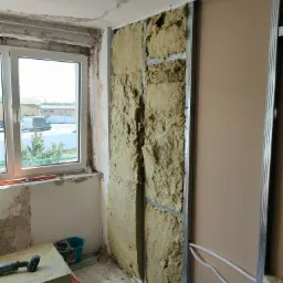 Ocieplanie ściany wełną mineralną i montaż płyt gipsowo-kartonowych na metalowym stelażu w trakcie remontu, widoczne przewody elektryczne, okno z widokiem na zewnątrz.