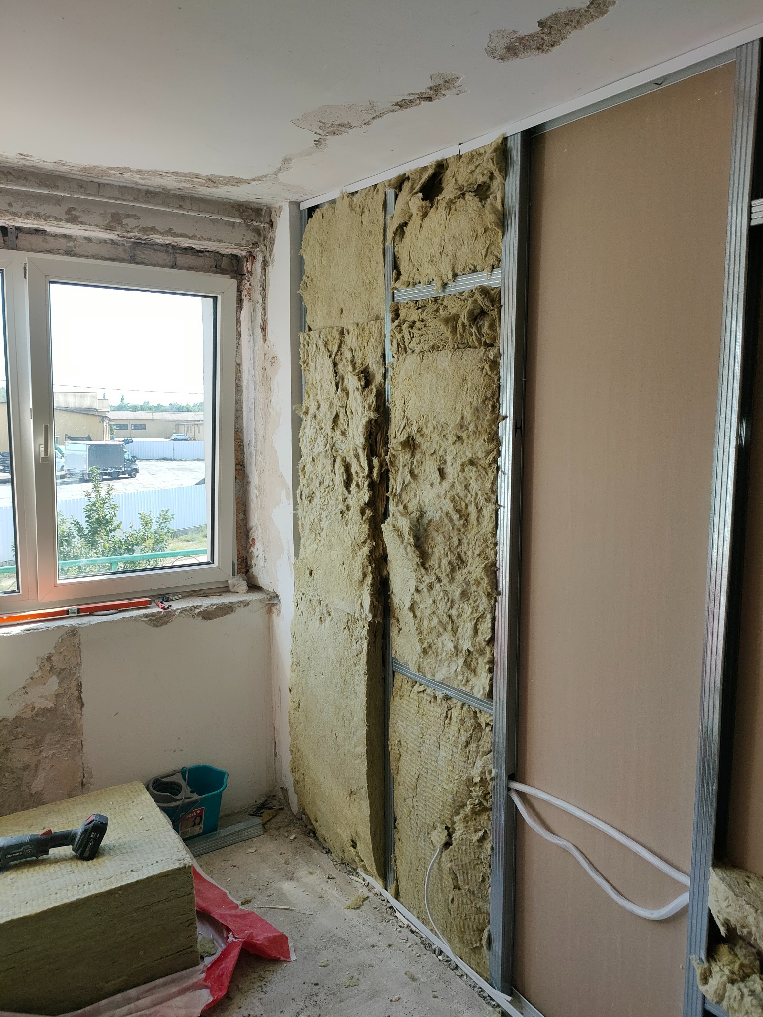 Ocieplanie ściany wełną mineralną i montaż płyt gipsowo-kartonowych na metalowym stelażu w trakcie remontu, widoczne przewody elektryczne, okno z widokiem na zewnątrz.