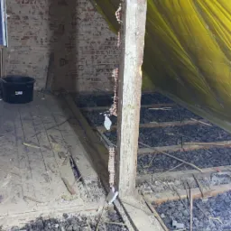 Poddasze w trakcie ocieplania: widoczne krokwie, żółta folia paroizolacyjna, rozsypany granulat izolacyjny oraz fragment odsłoniętej ceglanej ściany.