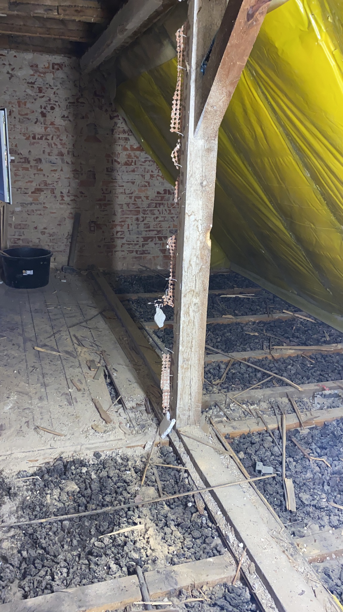 Poddasze w trakcie ocieplania: widoczne krokwie, żółta folia paroizolacyjna, rozsypany granulat izolacyjny oraz fragment odsłoniętej ceglanej ściany.