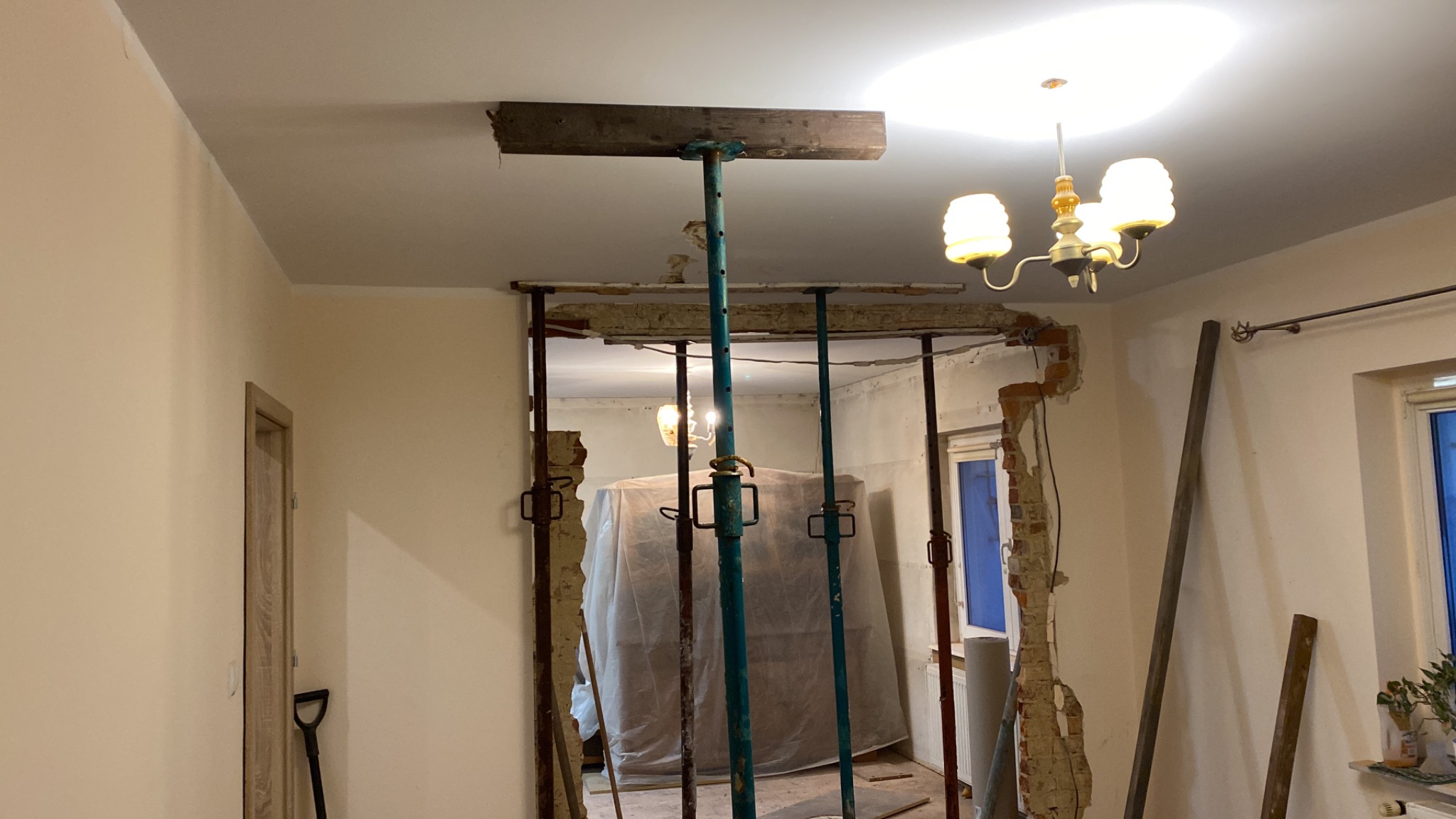 Wnętrze w trakcie remontu: wyburzona ściana podparta stemplami budowlanymi, odsłonięta cegła, widoczny gruz i zabezpieczone meble w tle.