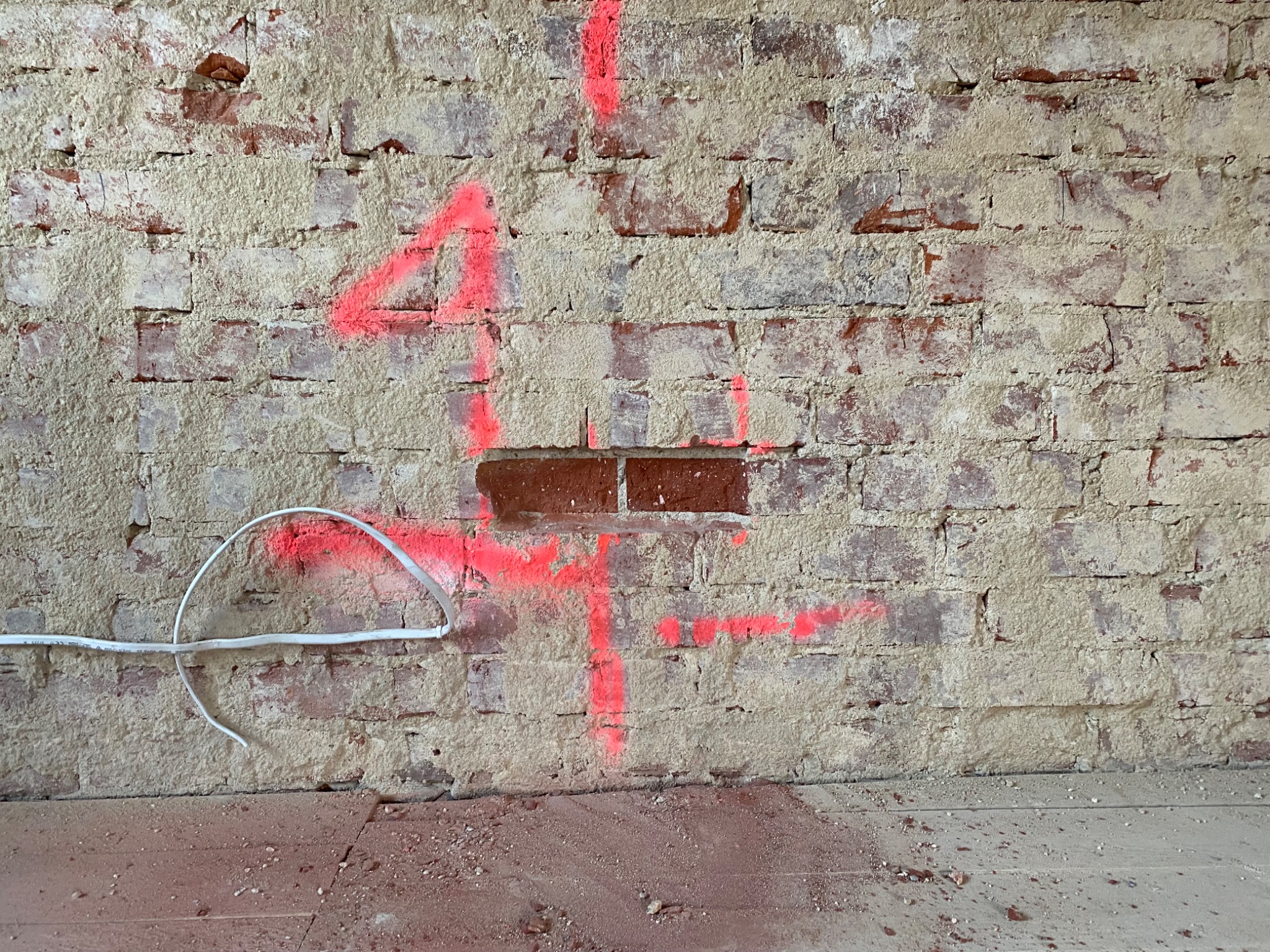 Ceglana ściana z pozostałościami zaprawy, zaznaczona neonowo-różową farbą w kształcie krzyża i cyfry '4', z wystającym przewodem elektrycznym i drobnymi odłamkami na podłodze.