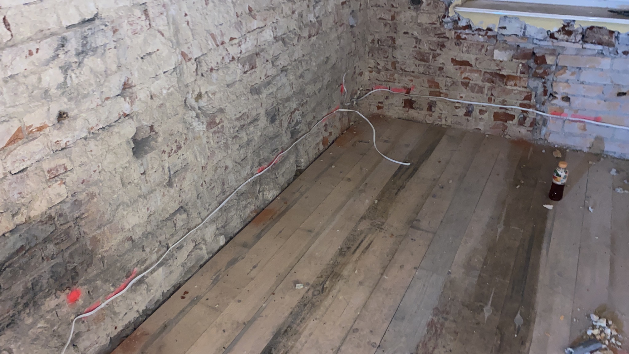 Pokój w trakcie remontu: odsłonięta cegła na ścianach z widocznymi śladami po instalacji elektrycznej, drewniana podłoga z butelką napoju w rogu.