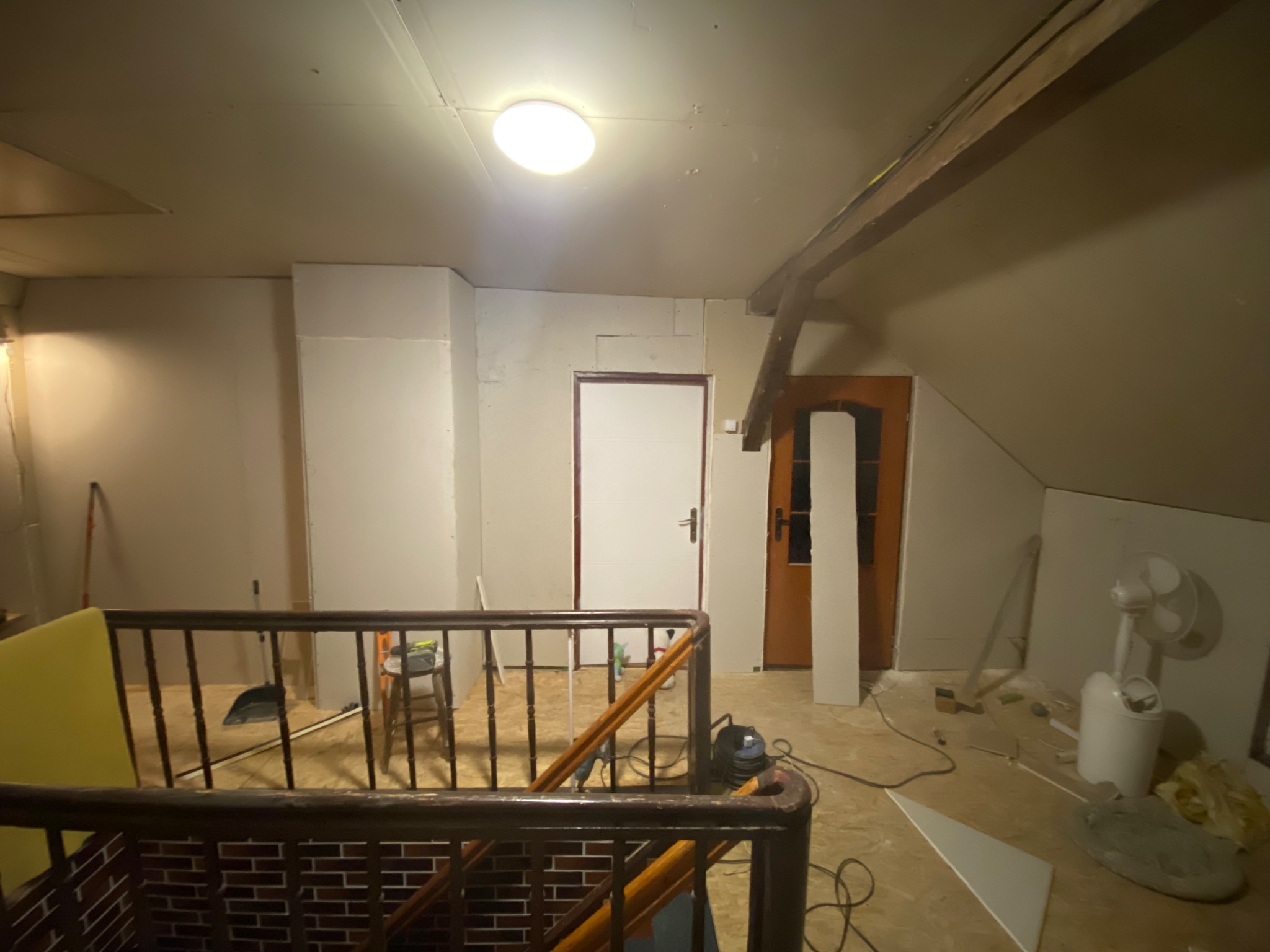 Poddasze w trakcie remontu: widok z góry na pomieszczenie z drewnianą belką, ścianami przygotowanymi do szpachlowania, narzędziami i wentylatorem.