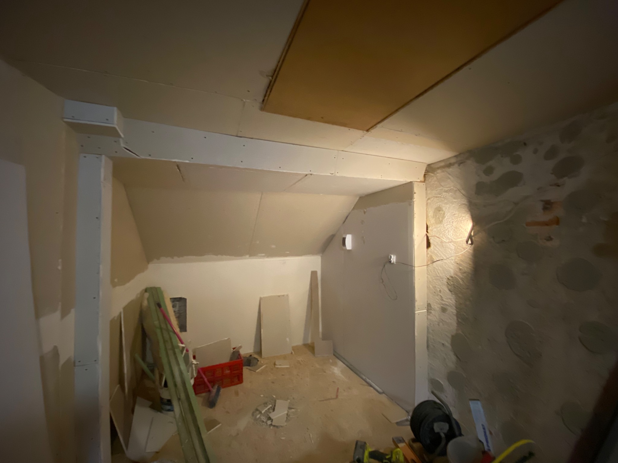 Poddasze w trakcie remontu, z widocznymi płytami kartonowo-gipsowymi na ścianach i suficie, fragmenty starego muru z pozostałościami tynku, narzędzia i materiały budowlane rozłożone na podłodze.