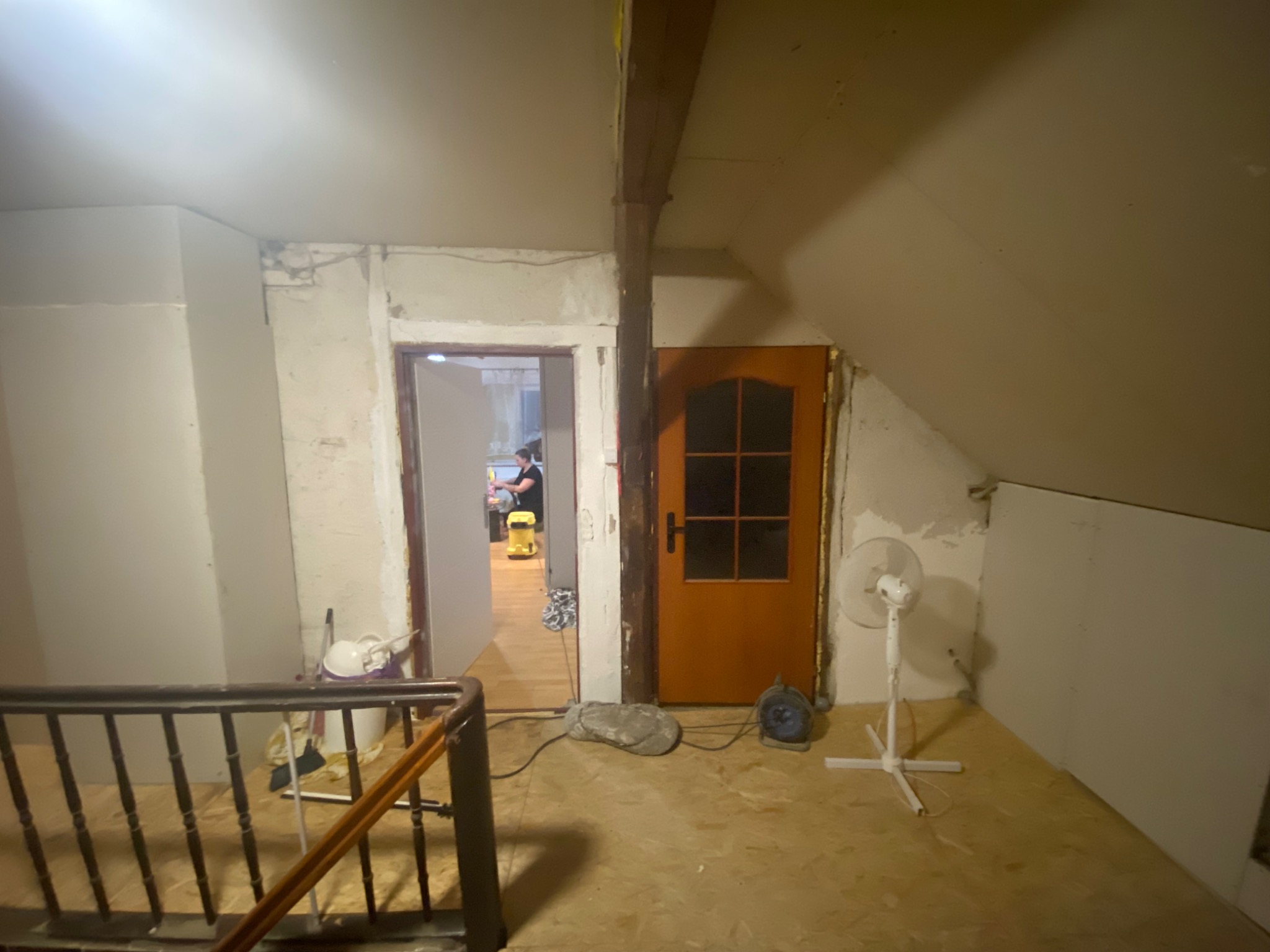 Poddasze w trakcie remontu: widoczny fragment schodów, surowe ściany przed wykończeniem, drewniana belka konstrukcyjna, wentylator i narzędzia malarskie. W tle widoczna osoba i odkurzacz.