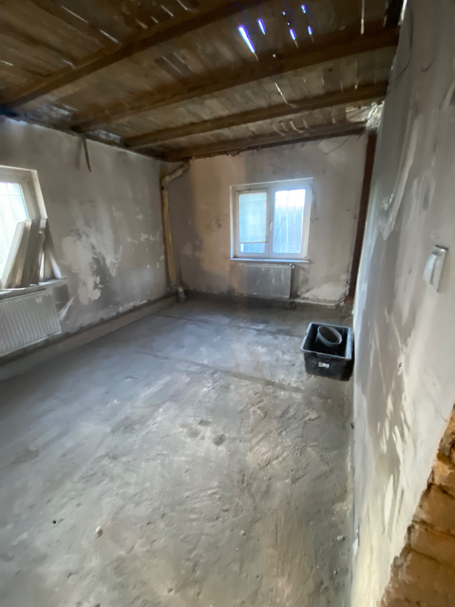 Pokój w trakcie remontu z surowym drewnianym stropem, ściany częściowo pokryte gładzią, widoczne okno i grzejnik, na podłodze resztki materiałów budowlanych i wiadro.