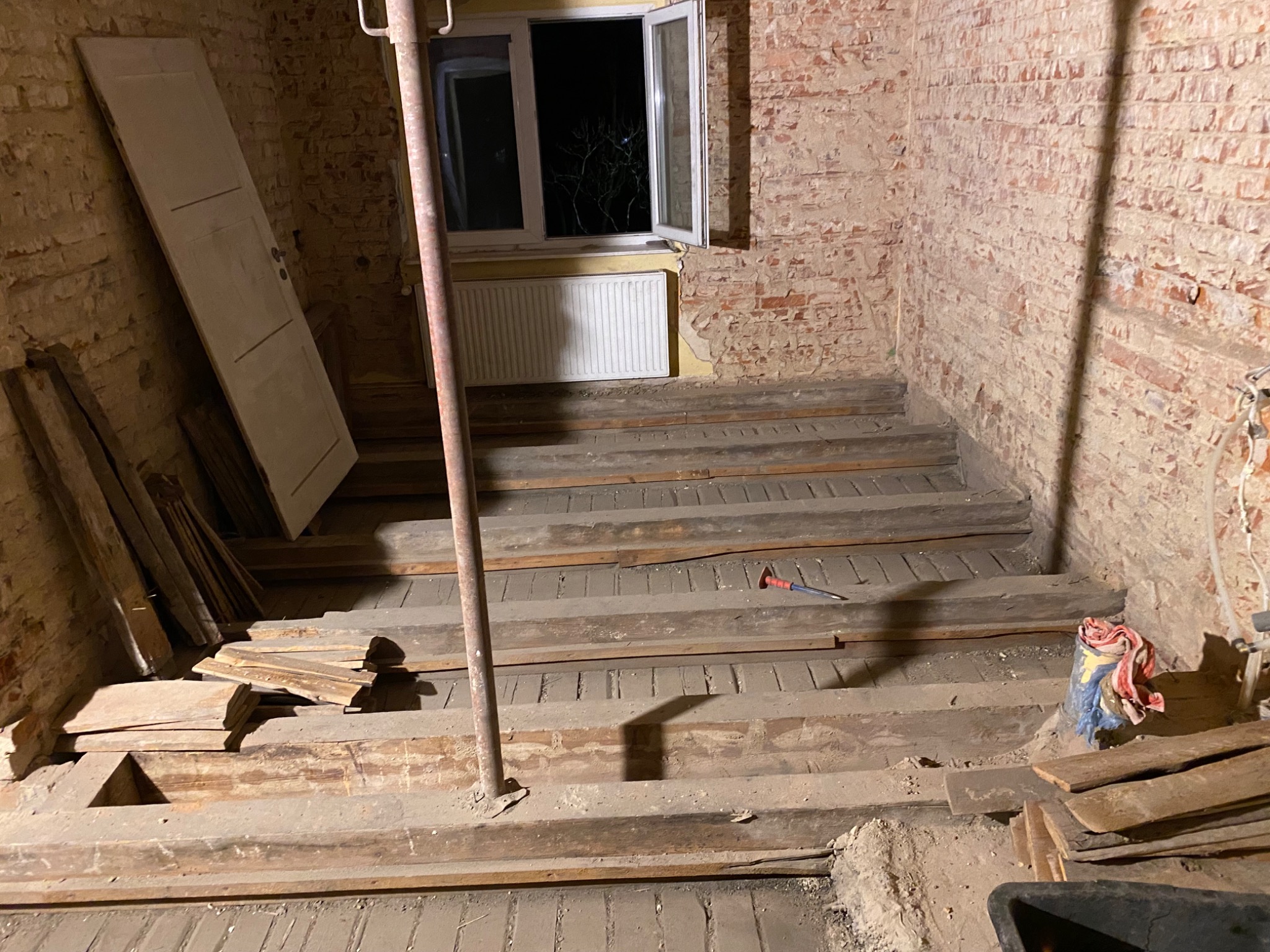 Wnętrze w trakcie remontu: surowe ceglane ściany z widocznymi śladami starej farby, drewniane stopnie schodów w trakcie budowy, podparte metalowym stemplem, okno z widokiem nocnego nieba, stara...