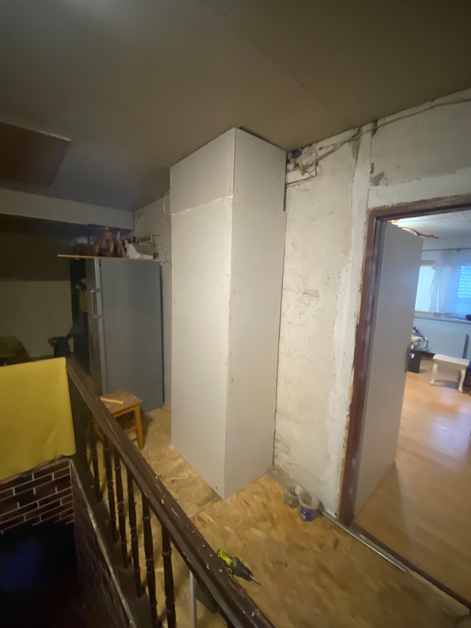 Montaż pionowej zabudowy z płyt gipsowo-kartonowych w pomieszczeniu z drewnianą podłogą i widoczną starą ścianą, w tle lodówka i schody.