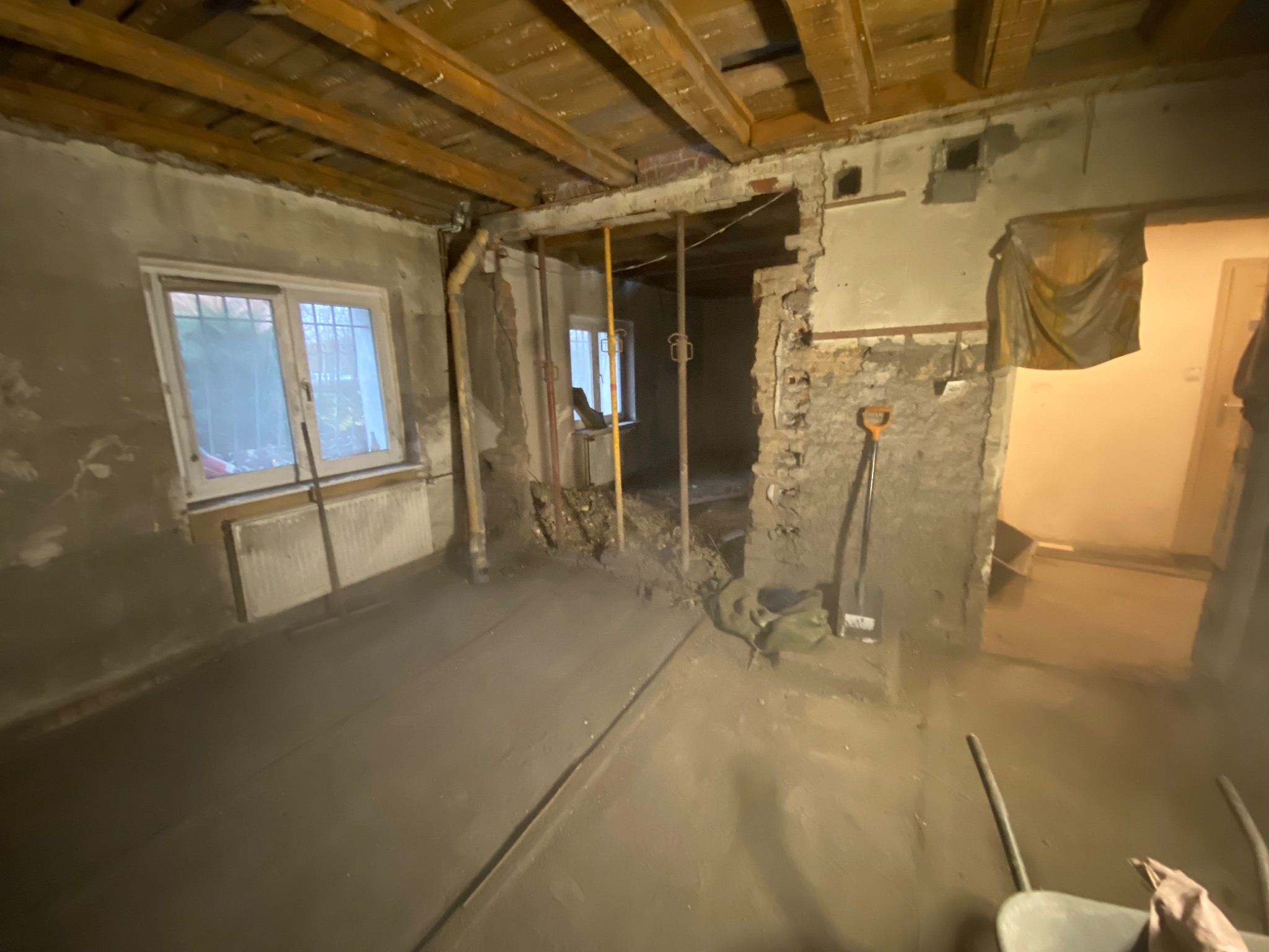 Wnętrze w trakcie remontu: świeżo wylana wylewka podłogowa, odsłonięte cegły, drewniany strop, widoczne narzędzia budowlane i podparte elementy konstrukcyjne.