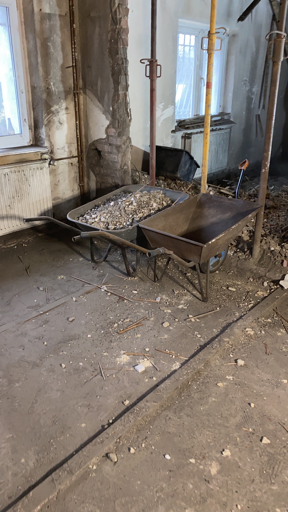Dwie taczki na podłodze w trakcie remontu, jedna wypełniona gruzem, widoczne odsłonięte fragmenty cegieł na ścianie i podpory budowlane.