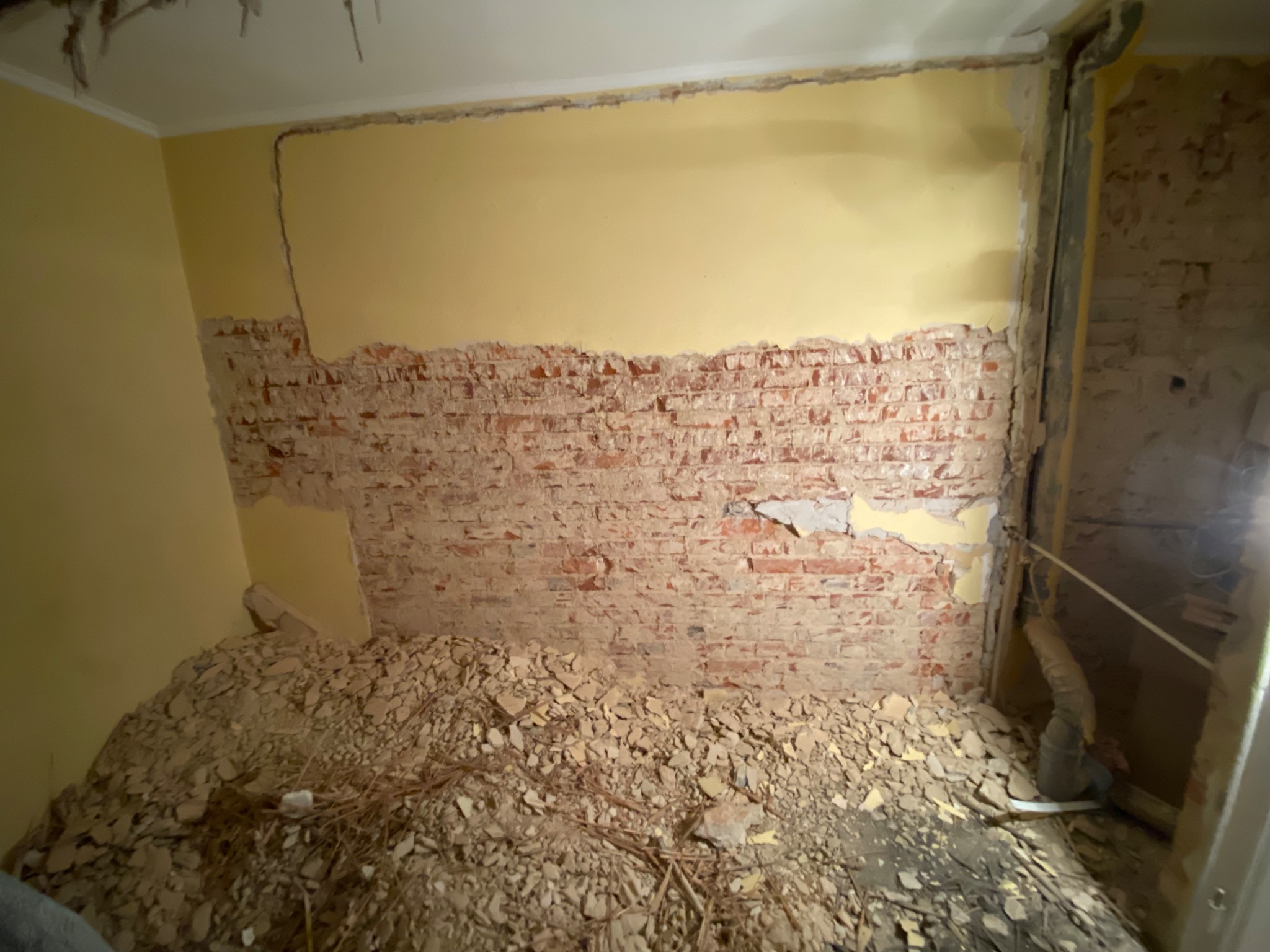 Fragment ściany z odsłoniętą czerwoną cegłą i resztkami żółtej farby, poniżej sterta gruzu i odpadów budowlanych.