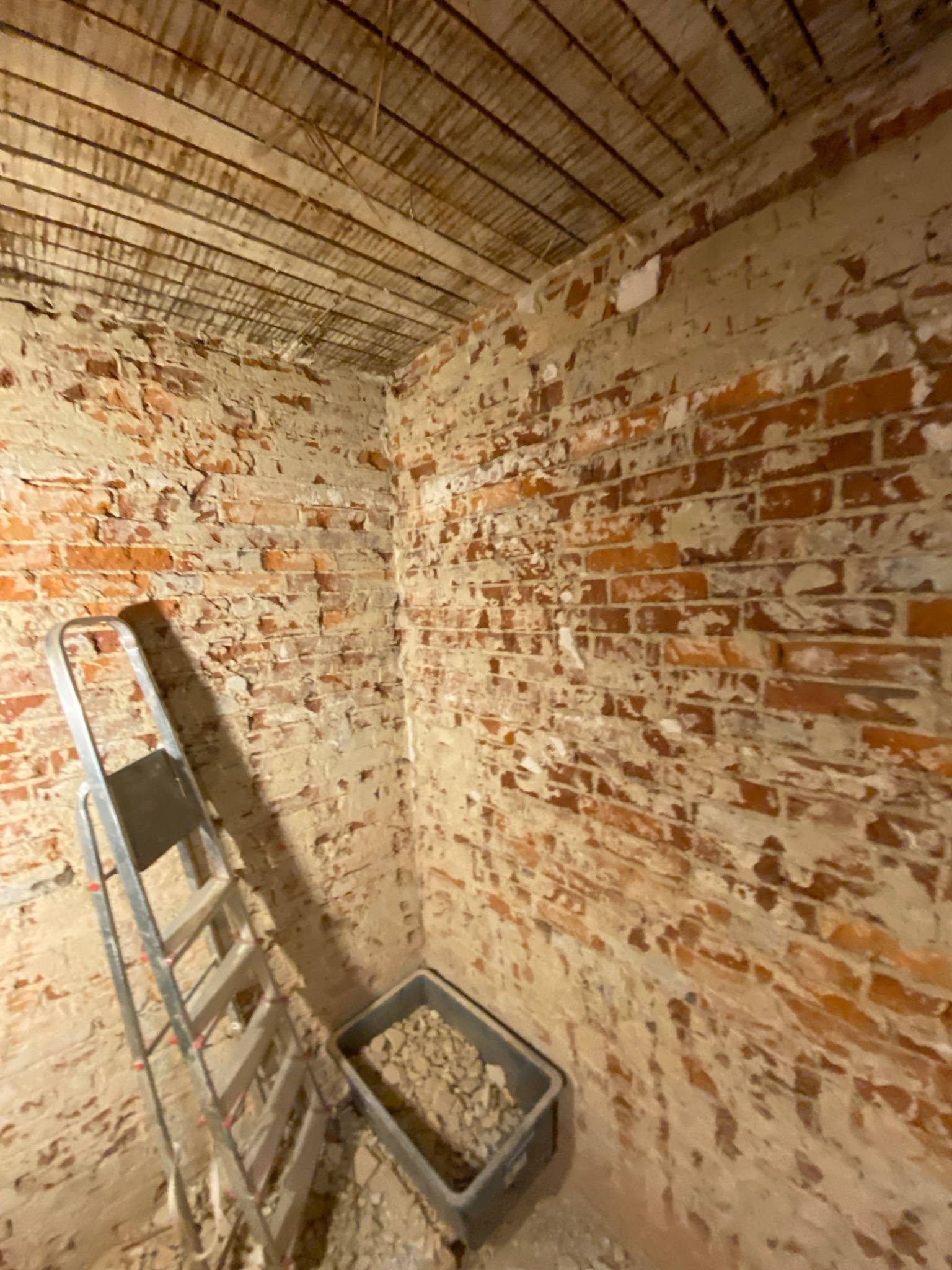 Surowe ceglane ściany w trakcie przygotowania do prac wykończeniowych, widoczny fragment drewnianego stropu, drabina aluminiowa oparta o ścianę oraz plastikowy pojemnik z gruzem.