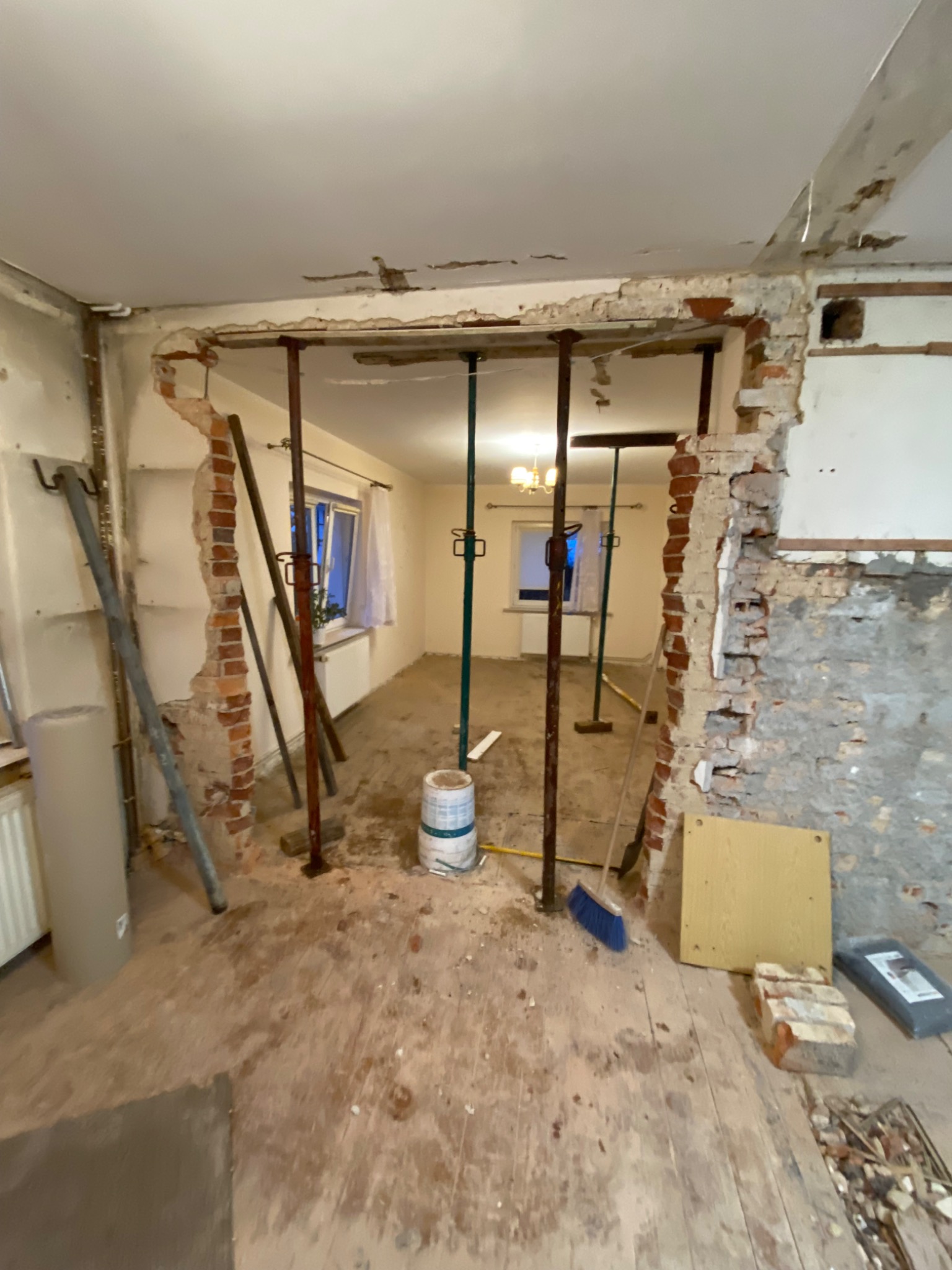 Demontaż ściany działowej z cegły, widoczne podpory stalowe, gruz, odkurzacz przemysłowy i odsłonięta podłoga w trakcie remontu.