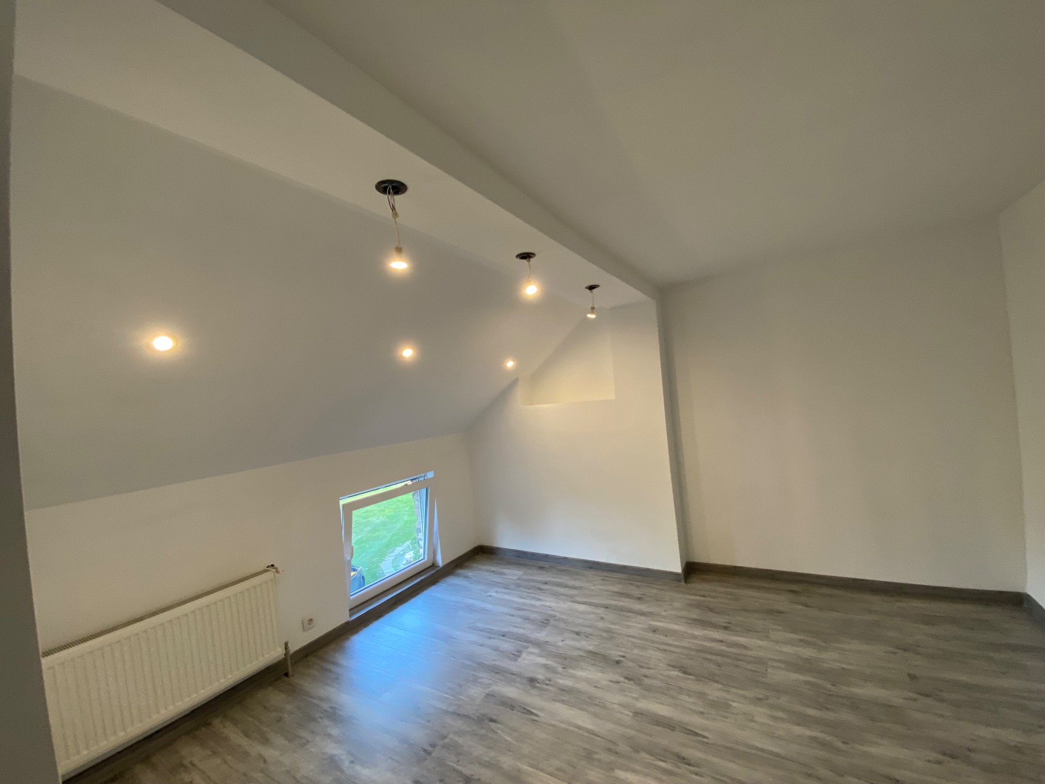 Poddasze po remoncie z panelami podłogowymi w odcieniach szarości, białymi ścianami i zainstalowanymi punktowymi lampami sufitowymi oraz grzejnikiem pod oknem.