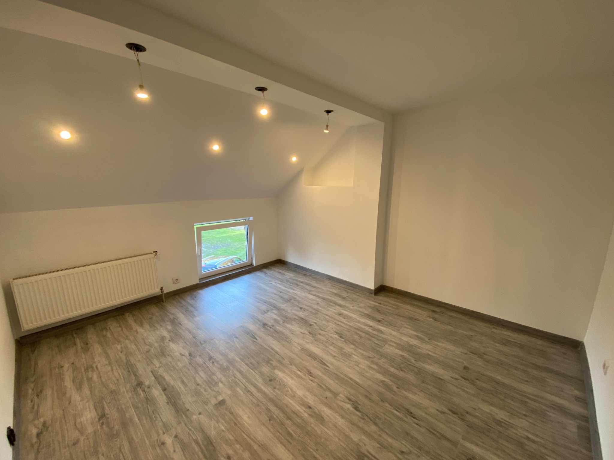 Poddasze po remoncie: świeżo pomalowane ściany na biało, panele podłogowe imitujące drewno, grzejnik pod oknem, kilka punktowych lamp sufitowych.