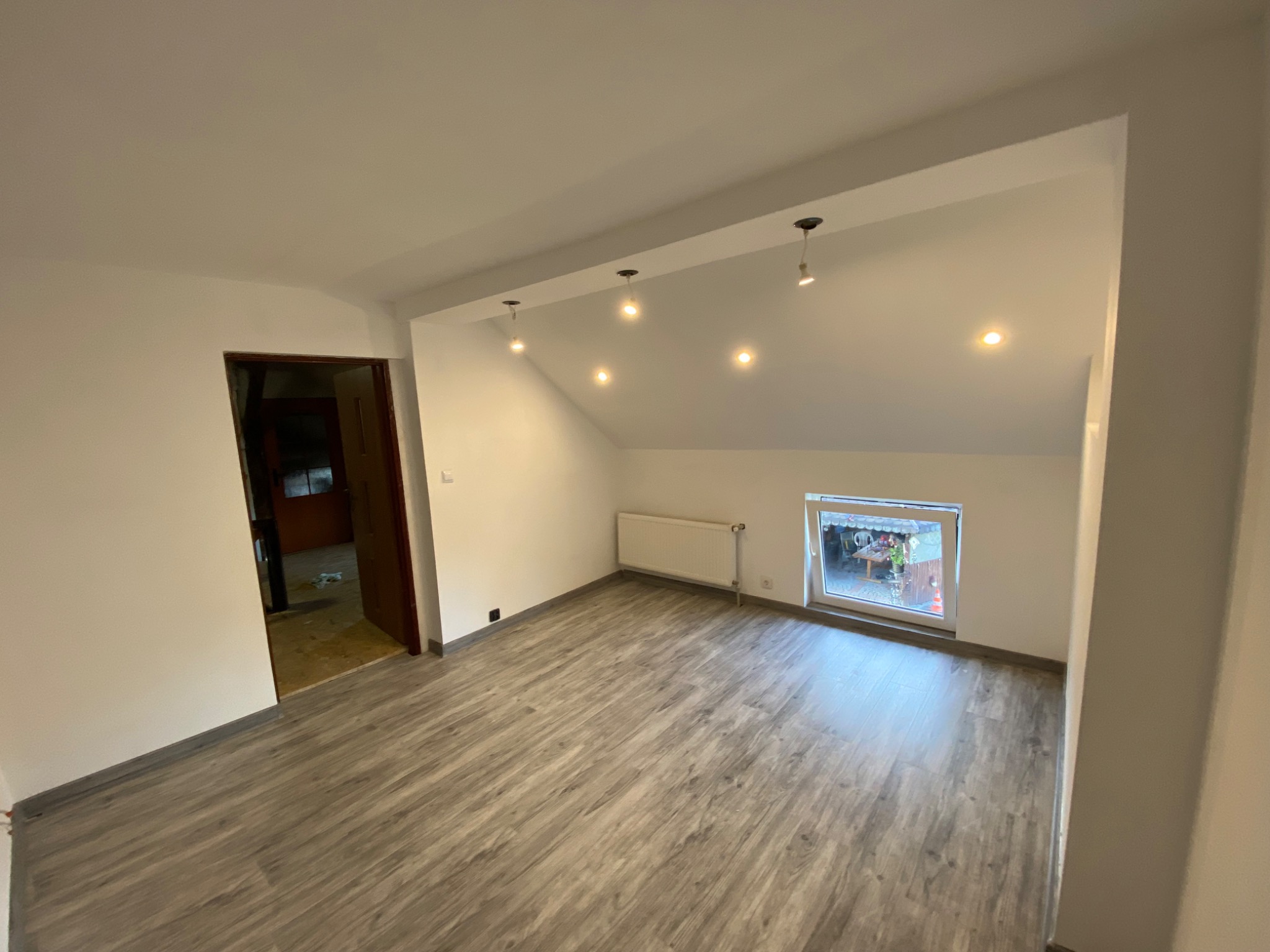 Poddasze po remoncie: świeżo ułożone panele podłogowe w odcieniach szarości, białe ściany z zamontowanymi punktowymi lampami sufitowymi, widok na okno i otwarte drzwi do sąsiedniego pomieszczenia.