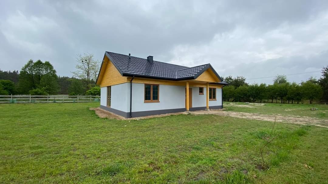 Nieduży, parterowy dom z szarą elewacją, drewnianą podbitką i ciemnym dachem z dachówki ceramicznej, otoczony trawnikiem i drzewami w pochmurny dzień.