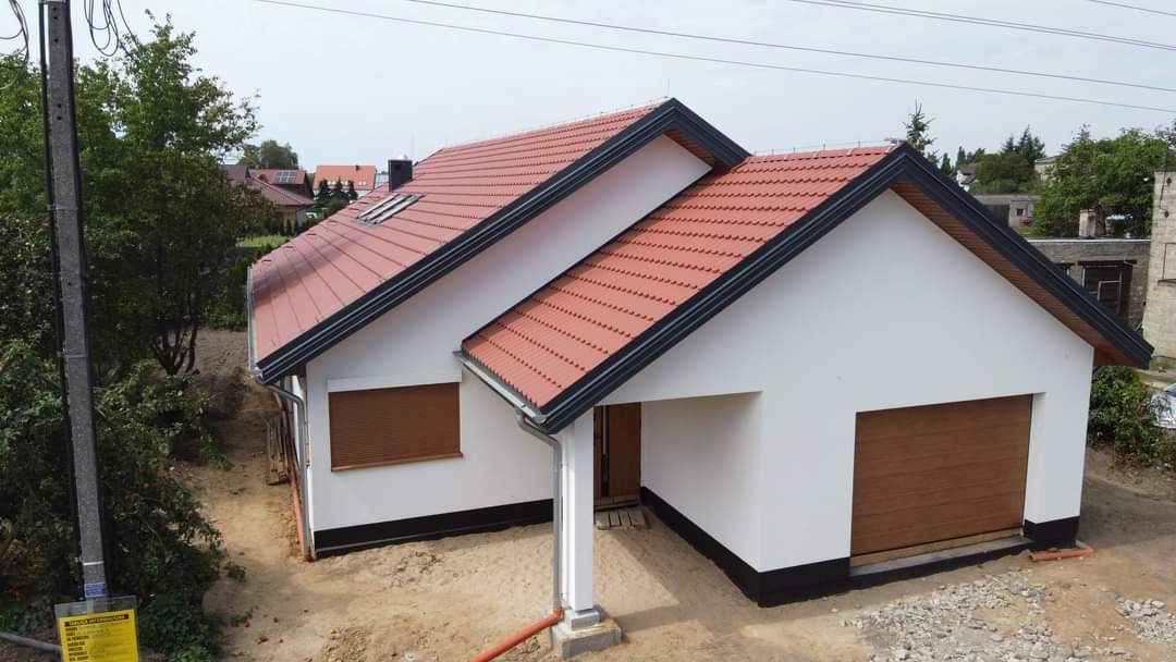 Nowo wybudowany dom jednorodzinny z czerwoną dachówką, białymi ścianami i brązową bramą garażową, widok z zewnątrz na niezagospodarowany teren.