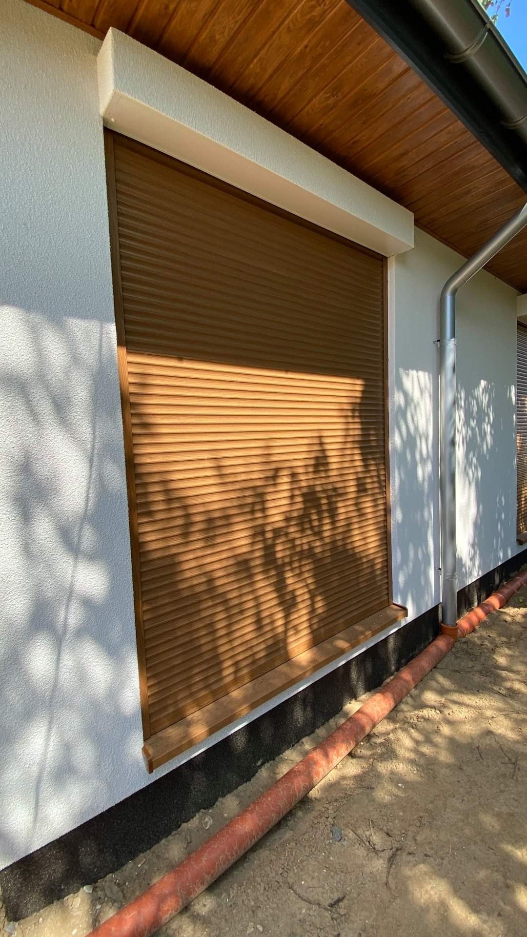 Zewnętrzna roleta okienna w kolorze drewna, zamontowana na otynkowanej elewacji budynku, z widocznym fragmentem orynnowania i instalacji podziemnej.