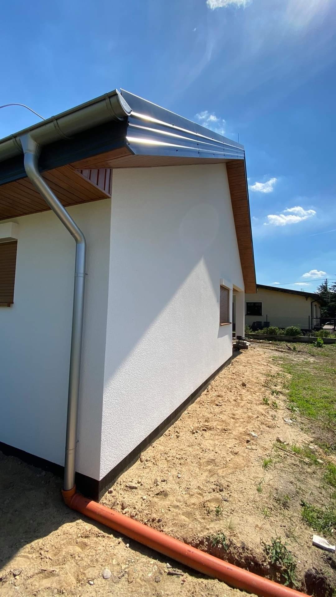 Boczny widok budynku z białą elewacją, metalową rynną i fragmentem dachu w słoneczny dzień. Widoczna pomarańczowa rura wzdłuż fundamentów i piaszczyste podłoże.