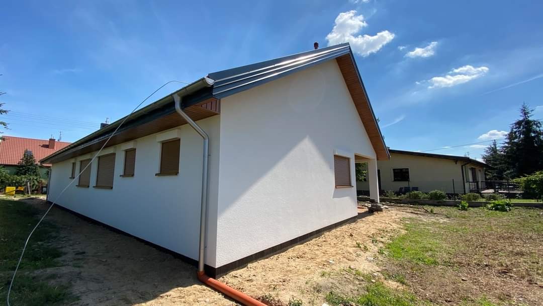 Nowoczesny, parterowy dom z białą elewacją i szarym dachem, widok z boku na tle błękitnego nieba z chmurami.