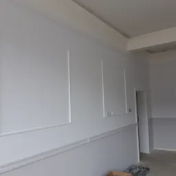 Świeżo pomalowane na biało ściany z dekoracyjnymi listwami, widoczne przygotowane profile podłogowe w kartonie i folii, surowy stan podłogi.