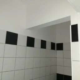 Ściana wyłożona białymi płytkami z poziomym pasem czarnych płytek w górnej części, widoczny fragment sufitu i narożnik ściany.