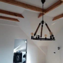 Wnętrze poddasza z drewnianymi belkami na suficie, nowym oświetleniem w postaci czarnego żyrandola z elementami liny i widokiem na łazienkę z pralkami.