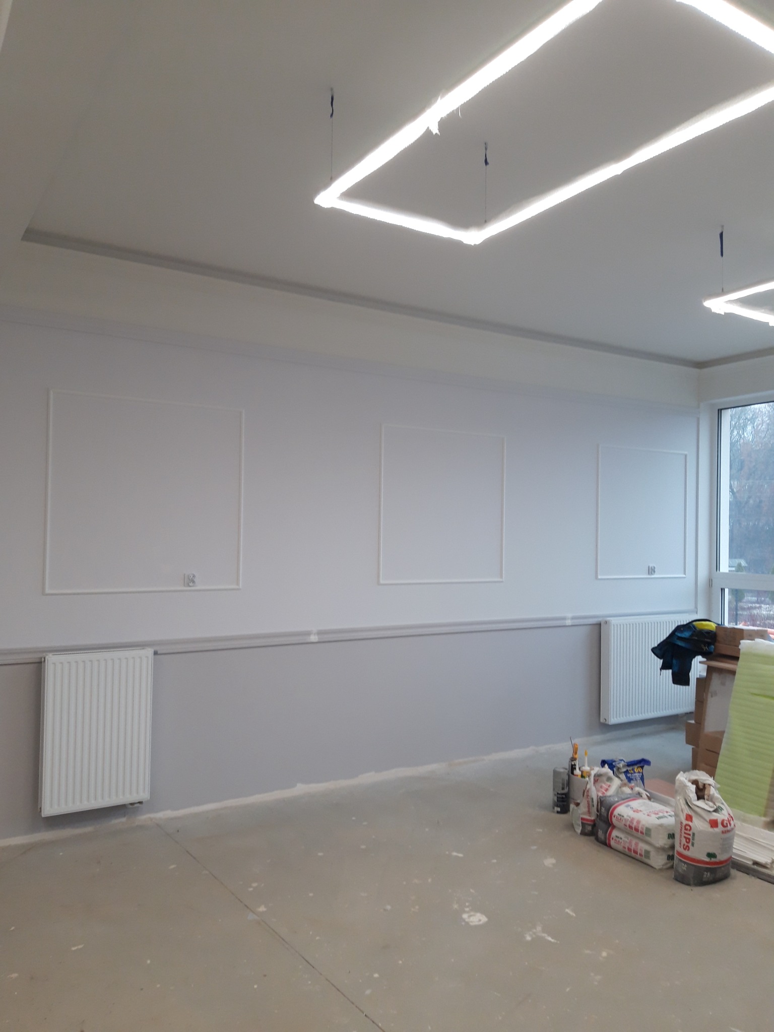 Wnętrze w trakcie prac wykończeniowych: szpachlowane ściany z dekoracyjnymi ramami, grzejniki, worki z gipsem na podłodze, oświetlenie sufitowe w kształcie kwadratów.