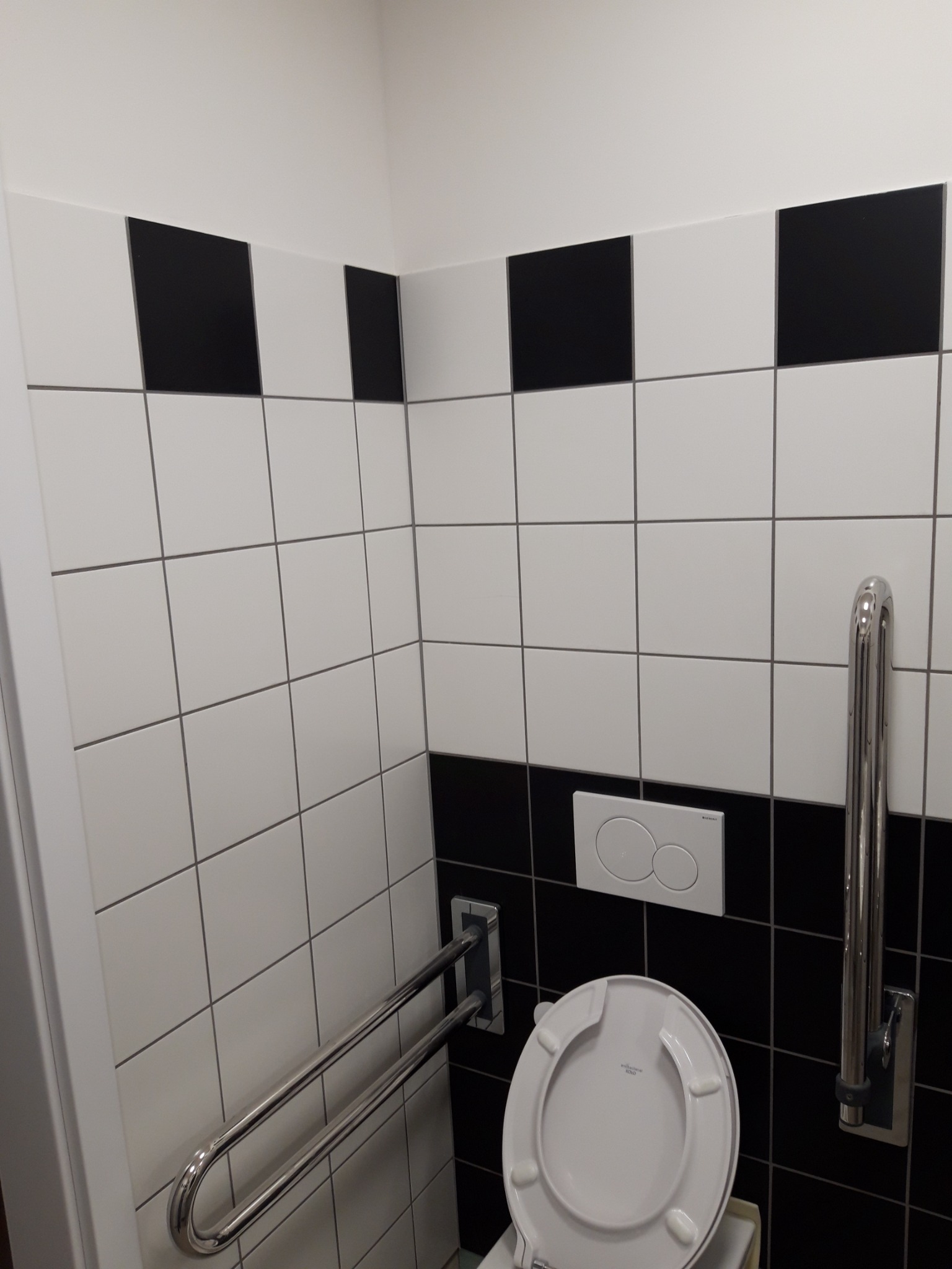 Wykończona łazienka z biało-czarnymi płytkami, uchwytem dla niepełnosprawnych i podwieszaną miską WC z przyciskiem spłukującym.