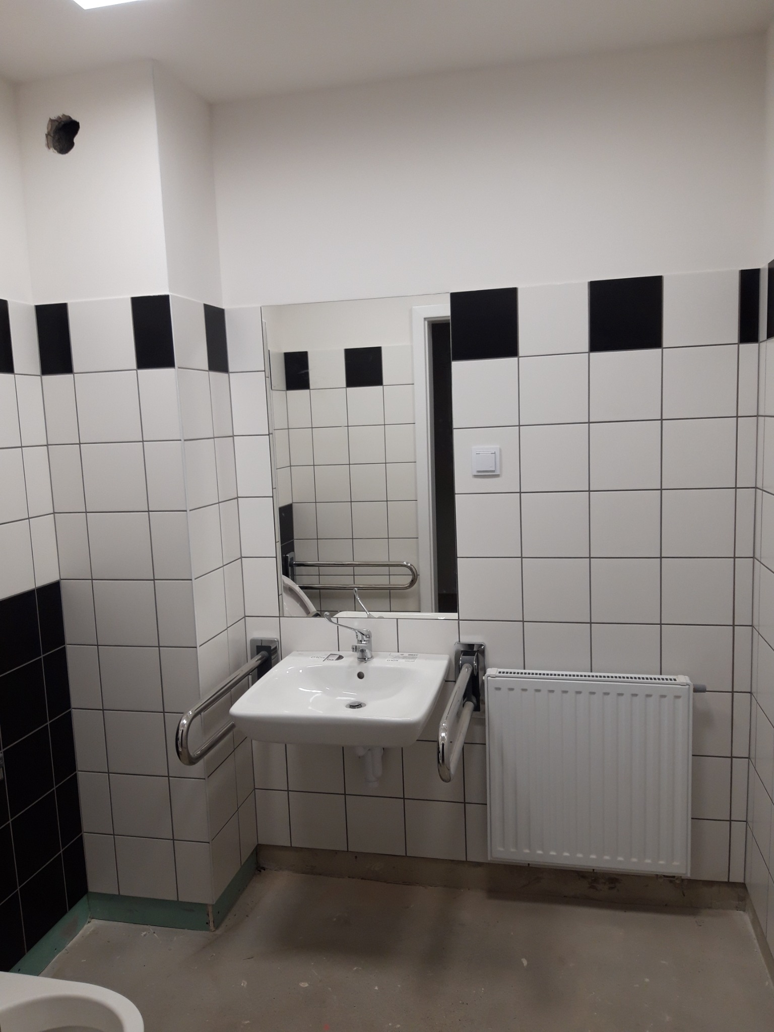 Łazienka publiczna z biało-czarnymi płytkami, umywalką dla niepełnosprawnych z poręczami i grzejnikiem, widoczny otwór w ścianie.