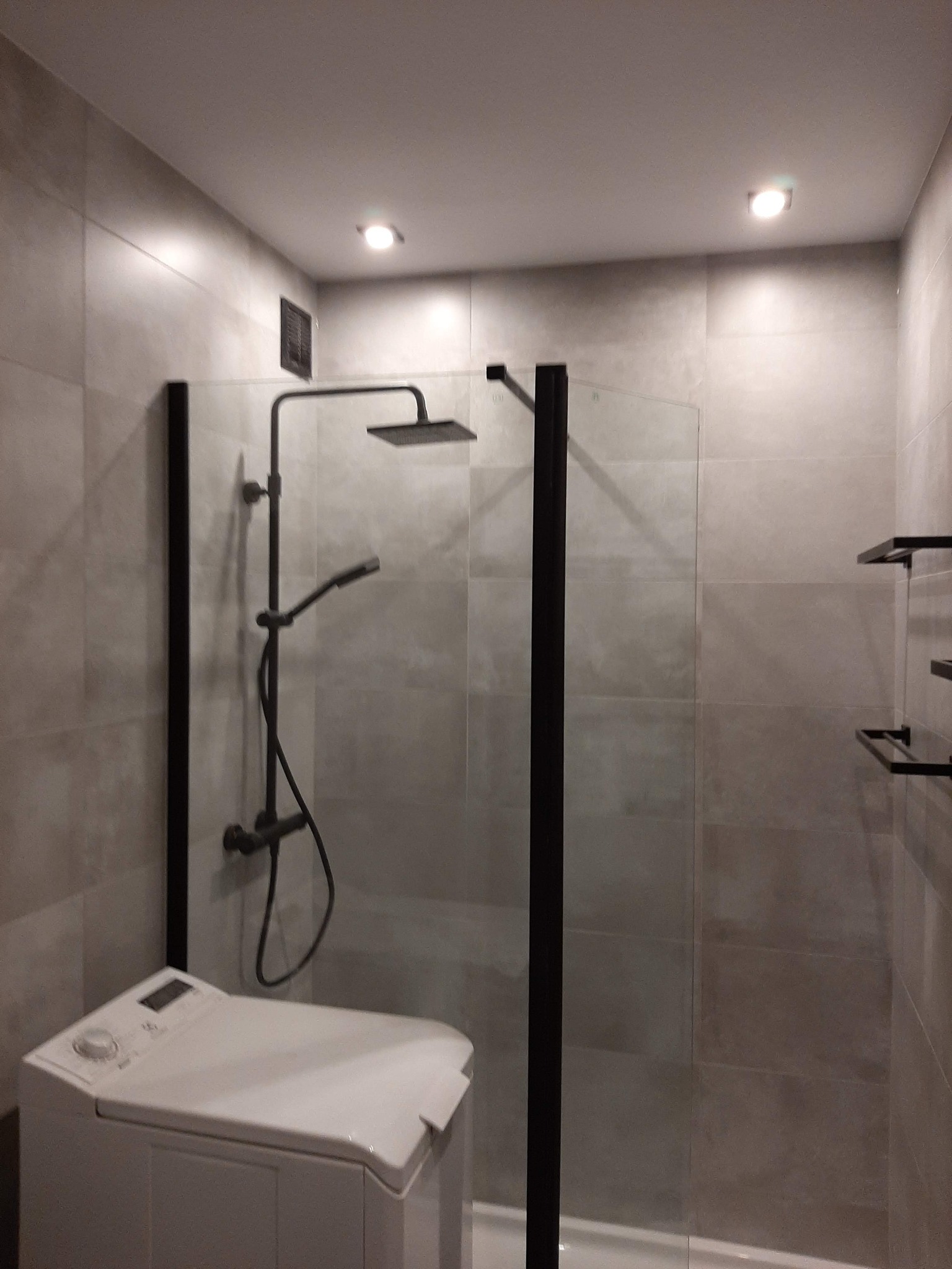 Nowoczesna łazienka z kabiną prysznicową typu walk-in z czarnymi ramami, deszczownicą i baterią w kolorze czarnym, obok pralka ładowana od góry, płytki w odcieniach szarości.