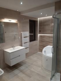 Jasna łazienka z beżowymi płytkami imitującymi drewno, okrągła wanna z kabiną prysznicową, biała szafka z umywalką i grzejnik drabinkowy, oświetlenie punktowe i listwa LED.