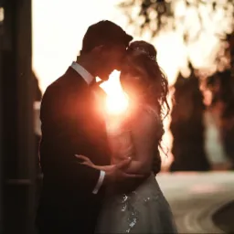 Para młoda w objęciach, silne, zachodzące słońce tworzy aureolę wokół ich sylwetek, subtelnie oświetlając suknię panny młodej zdobioną srebrnymi aplikacjami.