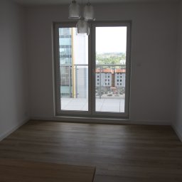 DAPO Firma Ogólnobudowlana - Pusty, jasny pokój z panelami podłogowymi i balkonem z widokiem na miasto, oświetlony wiszącą lampą.