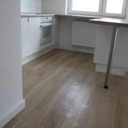 DAPO Firma Ogólnobudowlana - Jasne panele podłogowe imitujące drewno ułożone w nowym mieszkaniu z białą zabudową kuchenną i stołem barowym na jednej nodze.