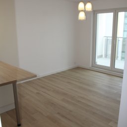 DAPO Firma Ogólnobudowlana - Jasne panele podłogowe imitujące drewno w pustym, nowym pomieszczeniu z białymi ścianami i potrójną lampą wiszącą, widok na balkon przez przeszklone drzwi.