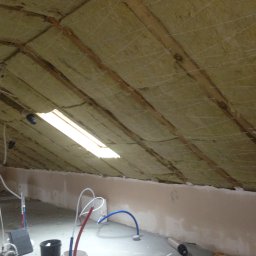 DAPO Firma Ogólnobudowlana - Montaż izolacji z wełny mineralnej na poddaszu, widoczne drewniane krokwie i siatka podtrzymująca, w tle pracownik w czerwonej koszulce umieszcza materiał izolacyjny.