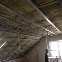 DAPO Firma Ogólnobudowlana - Poddasze w trakcie ocieplania wełną mineralną, widoczne metalowe profile konstrukcyjne pod płytę karton-gips, okno dachowe i fragment ściany szczytowej.