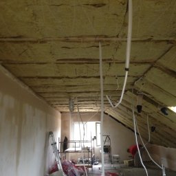 DAPO Firma Ogólnobudowlana - Poddasze w trakcie prac izolacyjnych: widoczna wełna mineralna przymocowana sznurkami do drewnianej konstrukcji dachu, białe rury instalacyjne, oraz fragment ściany z widocznymi śladami po...