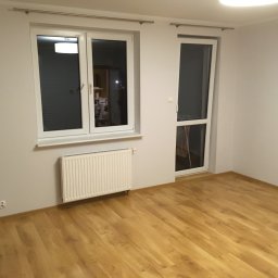 DAPO Firma Ogólnobudowlana - Pusty pokój z panelami podłogowymi w odcieniach ciepłego drewna, białymi ścianami, grzejnikiem pod oknem i drzwiami balkonowymi.