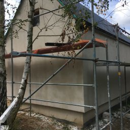 DAPO Firma Ogólnobudowlana - Budynek jednorodzinny w trakcie prac wykończeniowych elewacji, widoczne rusztowania, świeżo nałożony tynk, fragment dachu z blachy oraz brzozy na pierwszym planie.