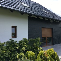 DAPO Firma Ogólnobudowlana - Nowoczesny dom z białą i grafitową elewacją, czarnym dachem z lukarnami i szarym tarasem, otoczony zielenią.