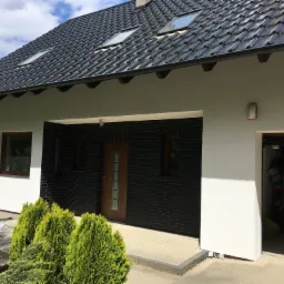 Wykończony dom jednorodzinny z czarną dachówką, białymi i czarnymi elewacjami, oraz widocznymi oknami dachowymi i zagospodarowanym terenem przed wejściem.