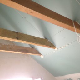 DAPO Firma Ogólnobudowlana - Poddasze w trakcie prac wykończeniowych: widoczne drewniane belki konstrukcyjne, płyty gipsowo-kartonowe zamontowane na skosach, szczeliny wypełnione pianką montażową oraz przewody elektryczne.